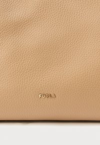 Beżowa skórzana torba o teksturowanej powierzchni. Na dole znajduje się złote, tłoczone logo "Furla", podkreślające minimalistyczny design.