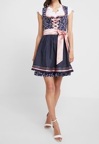 Kobieta w granatowej sukience dirndl w białe kwiatowe wzory, z różową satynową koronką na dekolcie, pasującą różową kokardą oraz białych sandałach na wysokim obcasie.