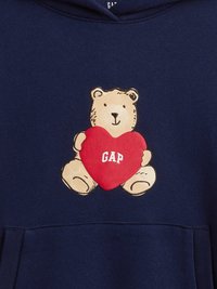 Tamsiai mėlynas džemperis su gaubtu, kuriame pavaizduotas lokys, laikantis raudoną širdį su užrašu „GAP“. Minkšta medžiaga, kanguro tipo kišenė ir gobtuvas su raišteliais.