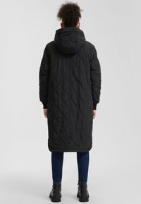 Zwarte gewatteerde lange jas met capuchon, met een diamanten patroon en elastische manchetten, gecombineerd met donkerblauwe jeans en zwarte enkellaarsjes.