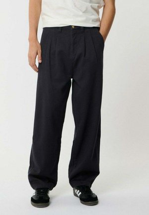 Person trägt lockere schwarze plissierte Hose, schwarz-weiße Sneakers und weißes Hemd, steht mit Händen in den Taschen.