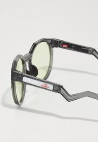 Graue transparente Sonnenbrille mit runden grünen Gläsern. Merkmale eines geometrischen Rahmen Designs und rotem "100 Thieves"-Logo am Bügel.