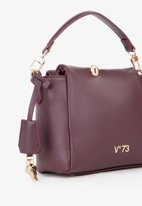 Bordeaux leren handtas met een gestructureerde afwerking, gouden hardware, een handvat aan de bovenkant en een sleutelhangeraccent. Bevat een opvallend merkembleem.