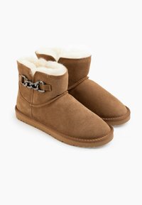 Botas de tobillo de ante marrón con un suave forro de piel blanca, que presentan un acento de cadena plateada y una suela de goma plana. Textura suave y forma redondeada.