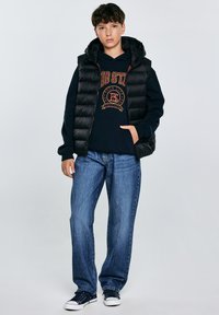 Chaleco puffer negro sobre una sudadera con capucha azul marino con un logo, combinado con jeans azules holgados y zapatillas oscuras con suelas blancas, que cuentan con capucha.