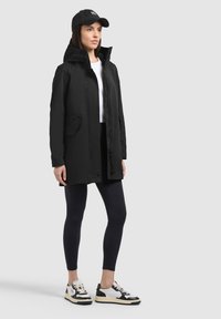 Žena v čiernom kabáte s kapucňou, čiernych legínach, bielom tričku, čiernej šiltovke a čierno-bielych teniskách stojaca na jednofarebnom pozadí.