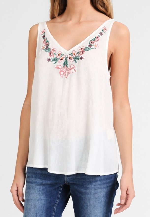 Femme portant un haut blanc sans manches à col en V avec une broderie florale rose et verte sur la poitrine, associé à un jean bleu.