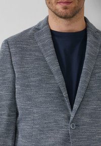 Grau strukturiertes Blazer mit einem Reverskragen, einem einzelnen Knopfverschluss und einem subtilen gewebten Muster, kombiniert mit einem dunkelblauen Rundhalsausschnitt-T-Shirt darunter.