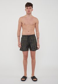 Pantaloni da bagno neri con ampie lettere gialle, vita elasticizzata e cordino interno. Il modello è in piedi a piedi nudi con sandali neri.