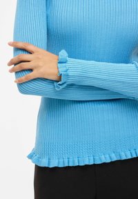 Personne portant un pull bleu côtelé avec des poignets et un ourlet à volants, les bras croisés sur le torse, associée à un pantalon noir.