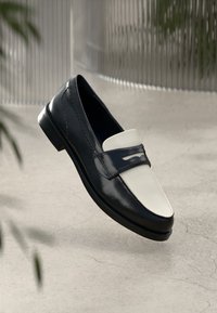 Mocassins de couro preto e branco suspensos acima de um chão de concreto, com plantas desfocadas e um fundo metálico estriado.