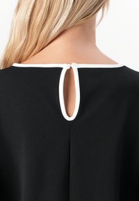 Femme blonde de dos portant un haut noir avec des bordures blanches et une fermeture à bouton en clé au niveau du col.