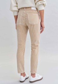 Beige Corduroyhose mit Slim Fit, vier Taschen und einem kleinen Rückentag, kombiniert mit weißen Sneakers und einem cremefarbenen Pullover.