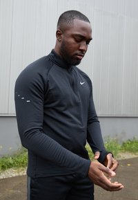 Crna dugorukava sportska majica s visokim ovratnikom, rebrastom teksturom i reflektirajućim detaljima na ramenima. Sadrži otvore za palčeve na rukavima.