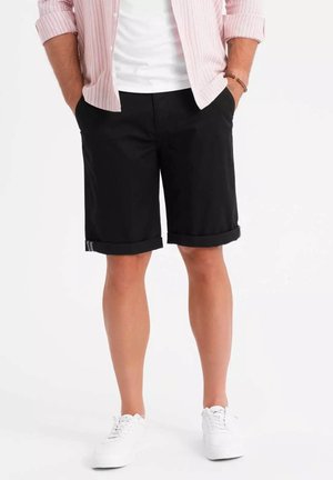Pantalones cortos negros con dobladillos enrollados, hechos de tela de algodón, con una textura suave y sutiles rayas grises en el dobladillo, combinados con zapatillas blancas.