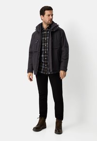 Ciemna ocieplana kurtka z kapturem, wieloma kieszeniami i zamkiem z przodu, noszona na niebieskiej koszuli w kratę, czarnych spodniach i brązowych butach.