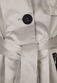 Trench coat de color gris claro con un acabado texturizado, que presenta un gran botón negro y un cinturón de tela, destacando su diseño entallado.