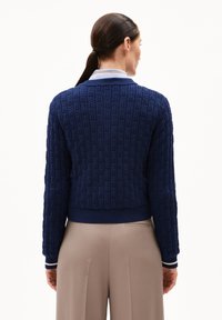 Dunkelblauer strukturierter Cardigan mit geripptem Saum und Bündchen. Mit einem gestrickten Muster. Getragen über hellbeigen Hosen.