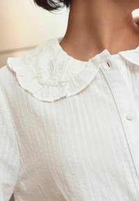 Chemise blanche avec un tissu texturé à rayures, un col froncé avec des motifs floraux brodés et une fermeture à boutons sur le devant.