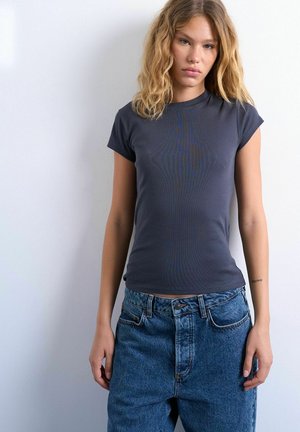 Young woman with shoulder-length blonde hair wears a fitted dark gray t-shirt and high-waisted blue jeans against a plain white background.