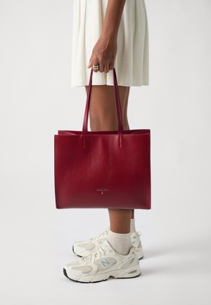Rød skinn tote bag med glatt tekstur, rektangulær form og to hendlene. Har en subtil logo på forsiden. Kombinert med hvite joggesko.