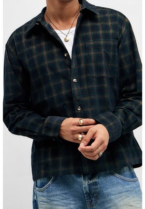 Person iført en mørkegrøn og brun ternet skjorte med knapper over en hvid t-shirt, lyseblå jeans og guld- og sølvringe på fingrene.
