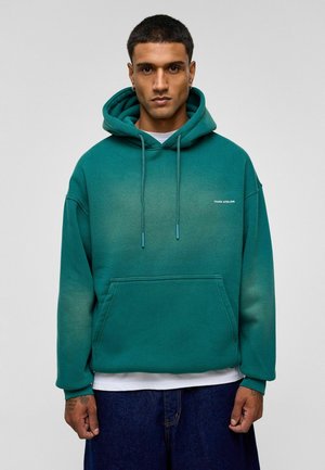 Groene hoodie met een zachte textuur, verstelbare trekkoorden, een zak aan de voorkant en een tonale logo. Draagt over een wit shirt, gecombineerd met donkere broeken.