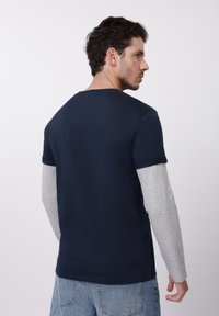 Tumši zils īsroku T-krekls virs pelēkas gari piedurkņu krekla. Kokvilnas audums ar klasisku apaļu apkakli un ikdienišķu pieguļošu stilizējumu.