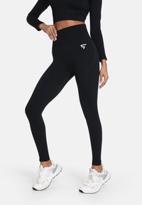 Čierne legíny s vysokým pásom, ribbovaným pásom a bielym logom, s hladkou textúrou a priliehavým dizajnom. Biele športové topánky.
