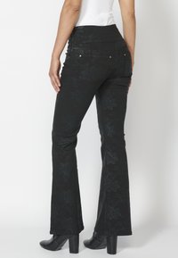 Jeans de campana con estampado floral negro, fabricados con un tejido texturizado, cintura alta, dos bolsillos traseros y costuras detalladas. Combinados con tacones negros.