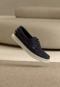 Μπλε ναυτικό παπούτσι από σουέτ με λευκή λαστιχένια σόλα, με αντιθετικές ραφές και κορδόνια, αιωρούμενο πάνω από μια υφή επιφάνεια.