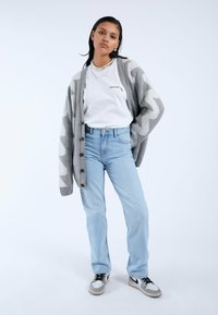 Cardigan gris con patrón en zigzag sobre una camiseta blanca, combinado con jeans azul claro y zapatillas grises. La modelo posa contra un fondo claro.