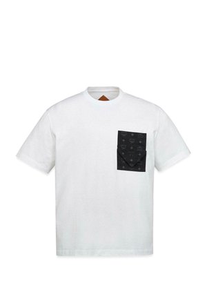 Jonge man die een wit T-shirt met een zwart borstzakje draagt over een patroon lange mouwen overhemd en losse zwarte broek, staand tegen een effen achtergrond.