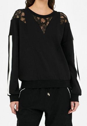 Sort bluse med lange ærmer med gennemsigtige blonde detaljer på skuldrene og øverste del af brystet, hvide striber langs ærmerne, kombineret med sorte bukser.