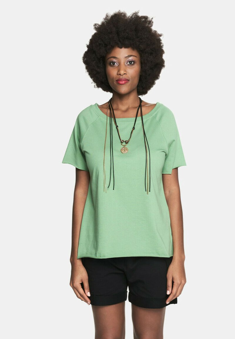 Jasnozielona bluzka z krótkim rękawem o luźnym kroju, z raglanowymi rękawami i szerokim dekoltem, w zestawie z czarnymi szortami i warstwowanymi naszyjnikami.