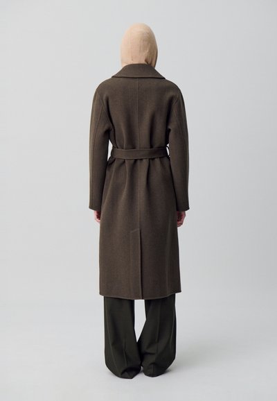 Cappotto lungo in lana marrone con vita a cintura, colletto ampio a punta e taglio dritto, abbinato a pantaloni a gamba larga verde oliva.