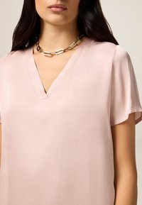 Blusa rosa chiaro a scollo a V con maniche corte, realizzata in tessuto morbido. Notevole collana d'oro con maglie intrecciate.