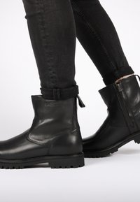 Botas de cuero negras hasta el tobillo con un acabado suave, suela texturizada en contraste, cremallera lateral y una pequeña lengüeta en el talón.
