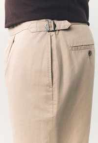 Beige Hosen mit flacher Vorderseite, Seitentaschen und einem Metallverschluss am Bund. Der Stoff wirkt glatt und leicht strukturiert.