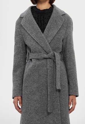 Personne portant un long manteau gris en laine avec un large col et une ceinture en tissu nouée à la taille sur un haut noir texturé.