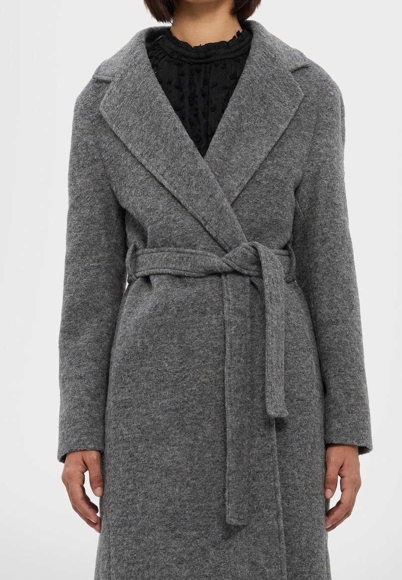 Personne portant un long manteau gris en laine avec un large col et une ceinture en tissu nouée à la taille sur un haut noir texturé.