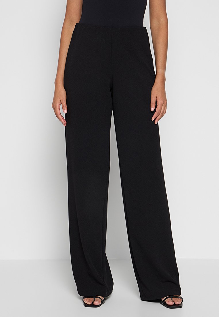 Femme portant un pantalon noir taille haute à jambes larges et des sandales noires à bout ouvert, se tenant contre un fond clair.