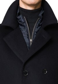 Podwójnie zapięty czarny płaszcz z szerokim kołnierzem, nałożony na ciemną kurtkę pikowaną z zamkiem błyskawicznym. Teksturowana wełniana tkanina i błyszczące akcenty.