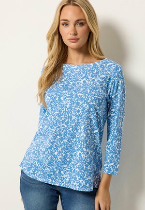 Frau mit langen blonden Haaren trägt ein blau-weiß gemustertes langärmeliges Oberteil und blaue Jeans, steht vor einer weißen Wand.