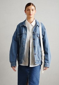 Blå denim oversized jacka med knappstängning, stora framfickor och avslappnad passform, bärs över en vit skjorta och en blå kjol.