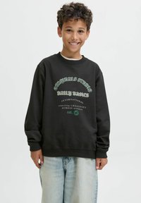 Felpa nera con scritta grafica verde, dotata di collo a giro e polsini a costine. Abbinata a pantaloni in denim chiaro, larghi.