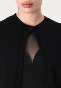 Μαύρο πλεκτό καρντάν με στρογγυλή λαιμόκοψη, κλείσιμο με ένα κουμπί και ριμπ επενδύσεις, τοποθετημένο πάνω από ένα σκούρο, διάφανο τοπ με V λαιμόκοψη.