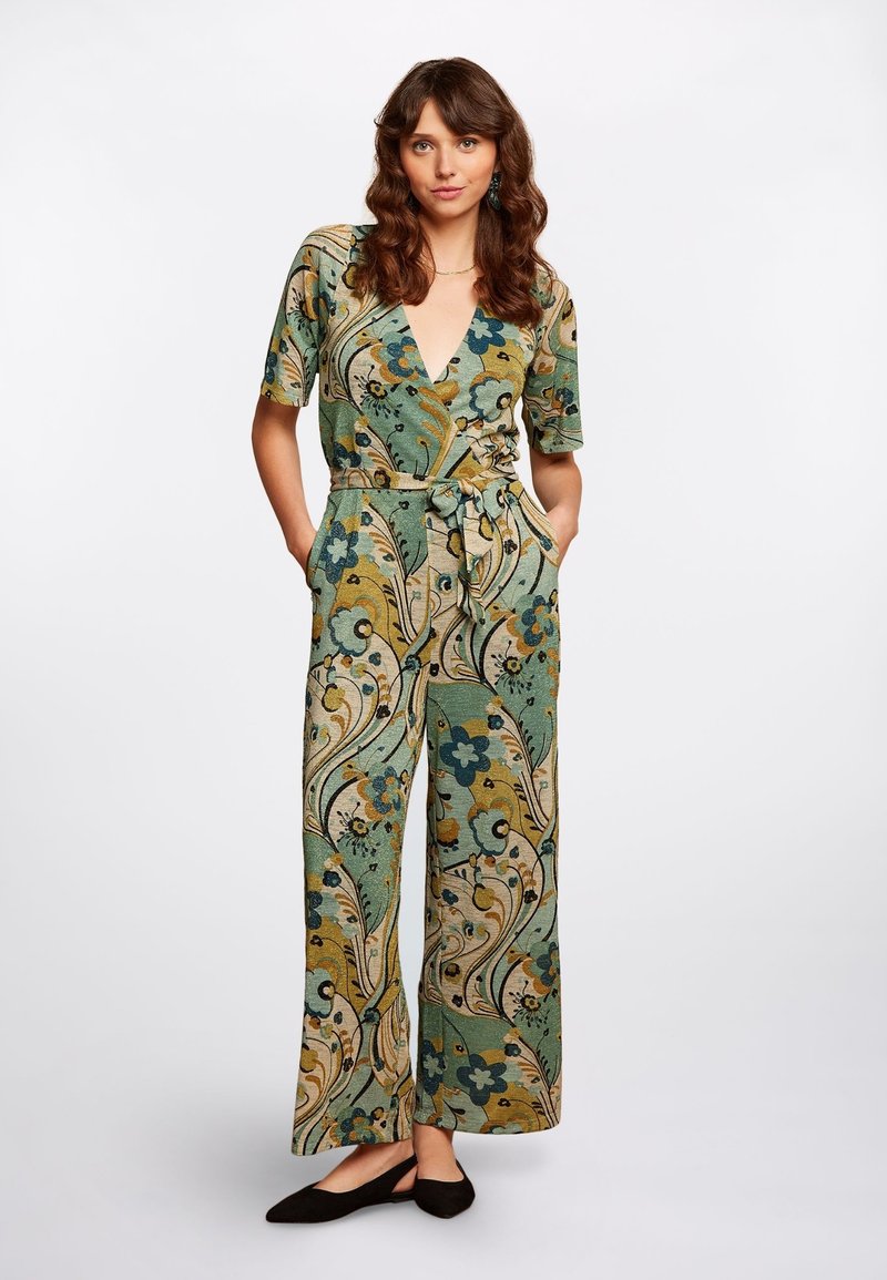 Mono floral en verde y beige, con un profundo escote en V, mangas cortas, cintura ceñida con cinturón y piernas anchas con patrones intrincados.