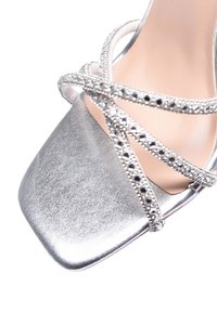 Scarpa argentata con tacco alto, caratterizzata da una punta quadrata, finitura metallica e cinturini incrociati decorati con piccoli gemme trasparenti.