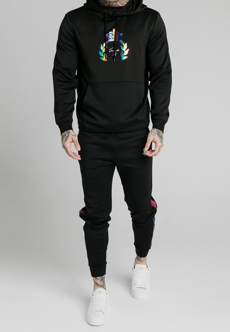 Czarna bluza z kapturem z kolorową grafiką na klatce piersiowej, połączona z czarnymi dresami z bocznymi akcentami w kolorze różowym i niebieskim. Białe sneakersy dopełniają stylizację.